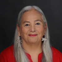 Dr. Denise Lajiimodiere, closed smile, wearing a bright red shirt, dangling dragonfly earrings with long grey hair
