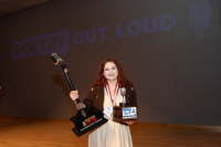 The ND 2024 State POL Champion, Circe Atkinson holding a large trophy, standing in front of a wall that reads Poetry Out Loud