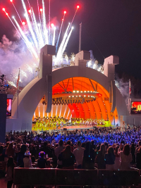 LA Philharmonic playing on the stage of the Hollywood Bowl, an outdoor ampitheatre with huge round backdrop to send sound forward, night photo with large crowd in seats and fireworks behind and above the stage