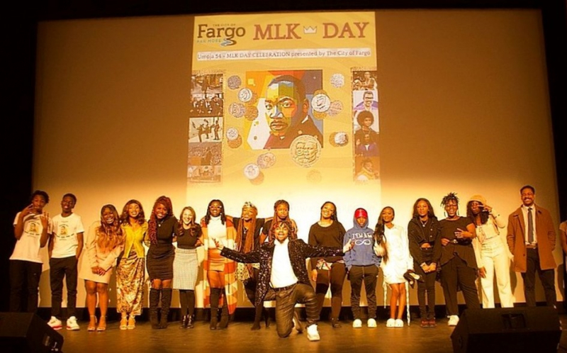 Frederick Edwards Jr. kneeling on stage with 16 black Americans and a Martin Luther King Jr. Day poster on the screen behind him behind him