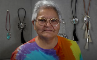 Head and shoulders of Nelda Schrupp with short grey hair, wearing glasses and tie dye t-shirt and her artistic Native jewelry hanging on the walls behind her
